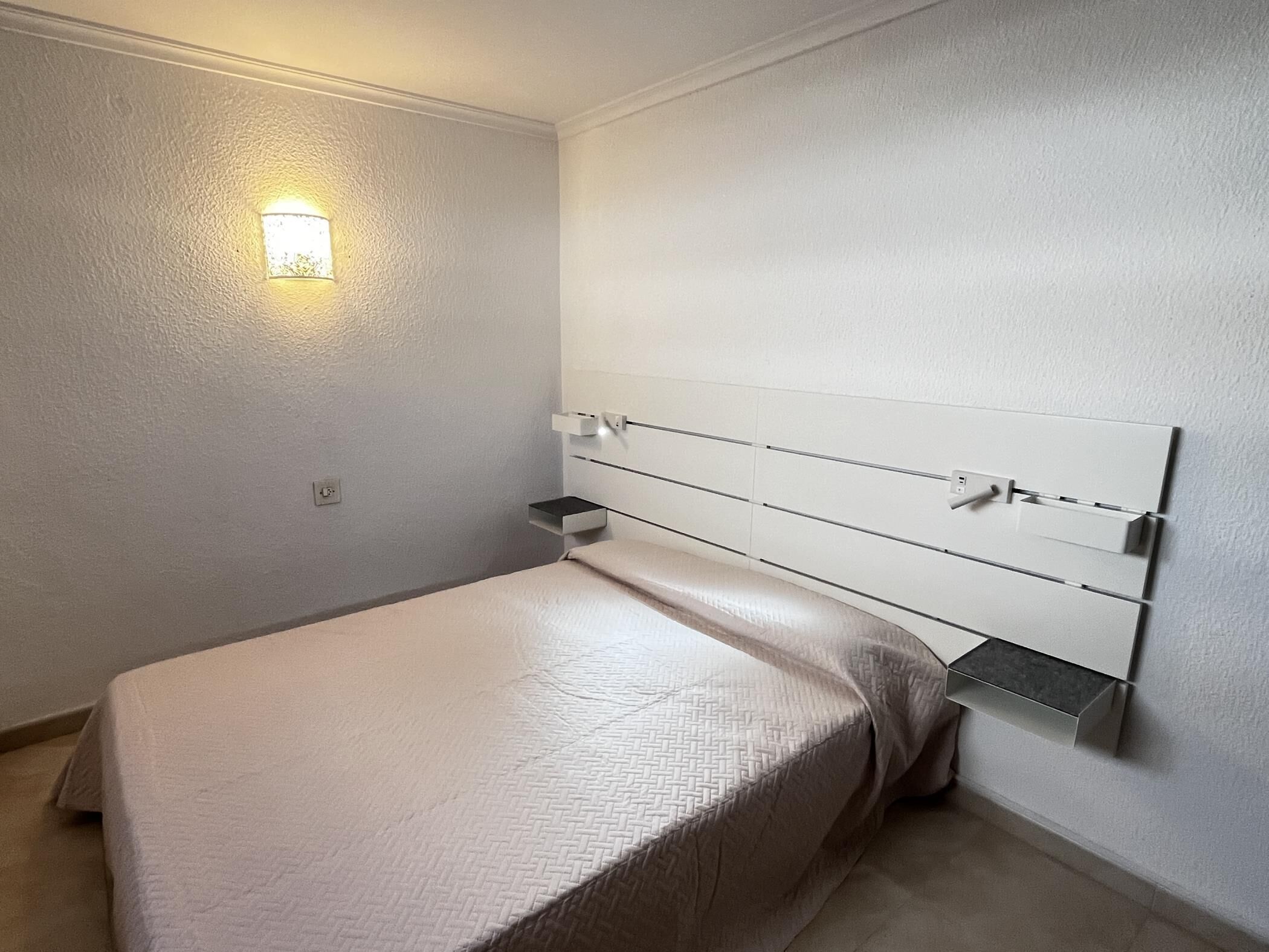 Un dormitori minimalista amb un llit, taules de nit i una làmpada de paret.