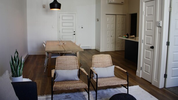 Uma sala de jantar com uma mesa de madeira e duas cadeiras de vime, uma luminária pendente e um vaso com planta.