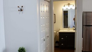 Um corredor com uma porta branca, uma pequena mesa com uma planta e um banheiro visível ao fundo.