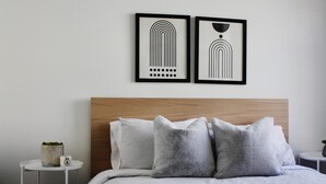Quarto bem arrumado com cabeceira de madeira, cama com roupa de cama cinza, duas mesas de cabeceira e duas obras de arte abstratas emolduradas na parede.