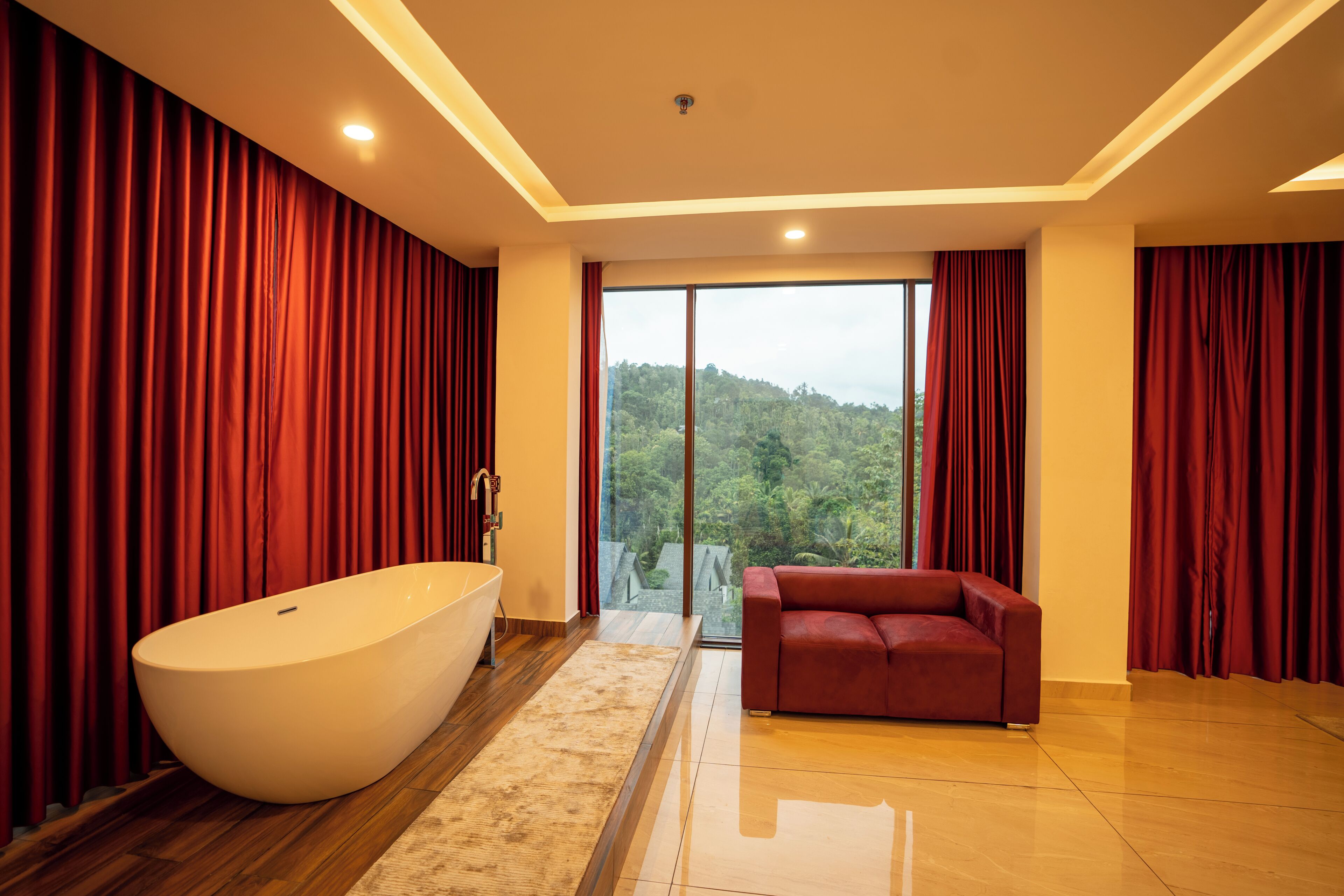 A modern bathroom with a freestanding bathtub, a red sofa, and a large window offering a view of a forested area.