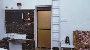 A compact living space with a TV mounted on a marble wall, a small shelf with decorative items, a refrigerator, a ladder, and a patterned sofa. - EASTERN GATE HOMESTAY (Rajnagar)