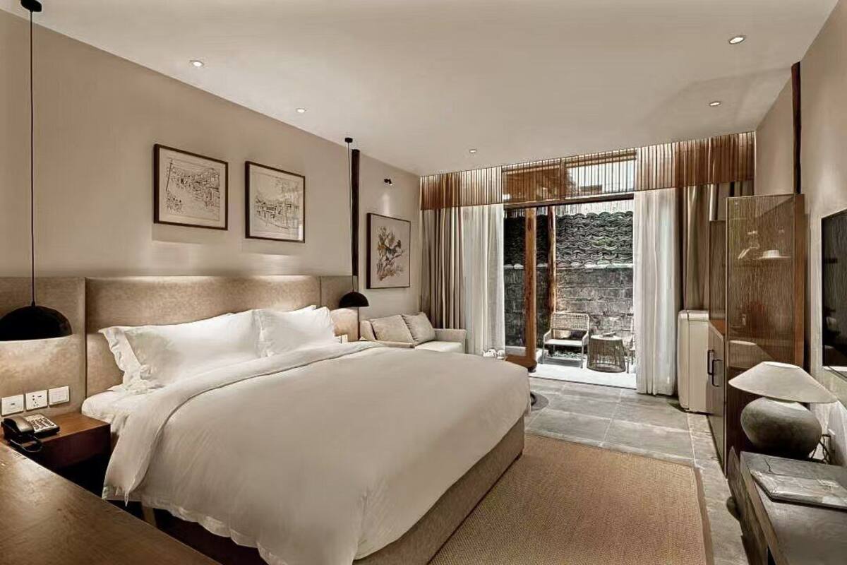 A modern hotel room with a large bed, a desk, a chair, and a TV. There is a balcony with a view of the city.