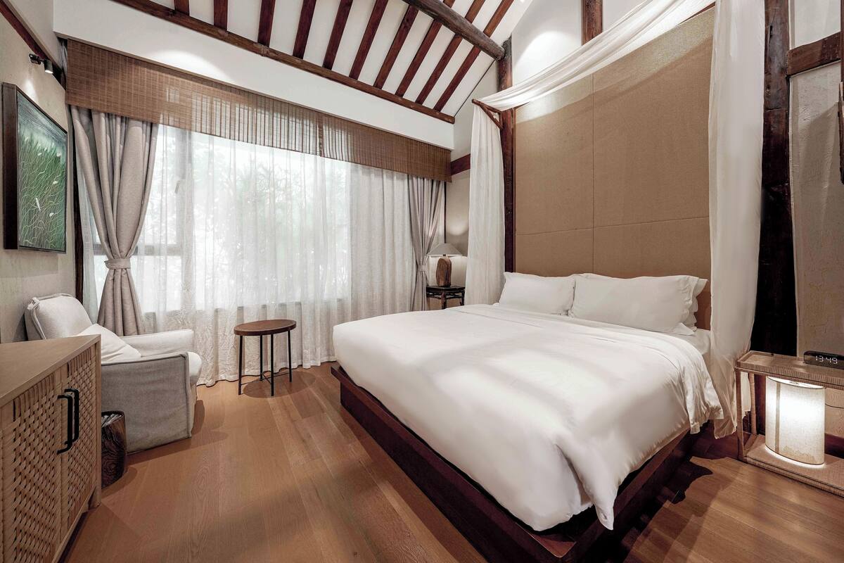 A spacious bedroom with a large bed, a bedside table with a lamp, a small side table, and a chair.