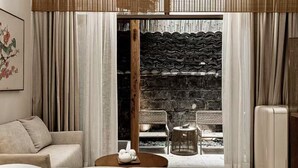 A hotel room with a bed, a sofa, a small round table with teapots, a wicker chair, and a view of a courtyard with a brick wall and a wooden beam. - Ancient Charm Inn Chengdu (Chengdu)