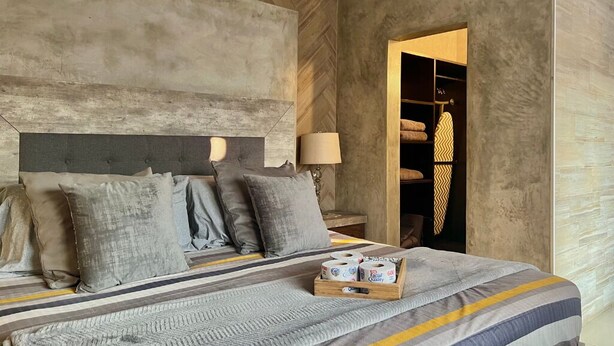 A bedroom with a bed, pillows, a bedside table with a lamp, and a box of toilet paper.