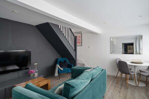 A modern living room with a large flat-screen TV, teal sofas, a wooden coffee table, a blue armchair, and a staircase with a black railing.