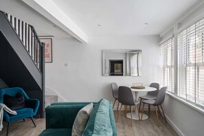 A modern living room with a teal sofa, a dining table with chairs, a staircase, and a large window with blinds. - Modern Cottage in Central Exeter (Devon)