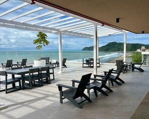 Ein überdachter Außenbereich mit Tischen und Stühlen, mit Blick auf einen Strand und das Meer.