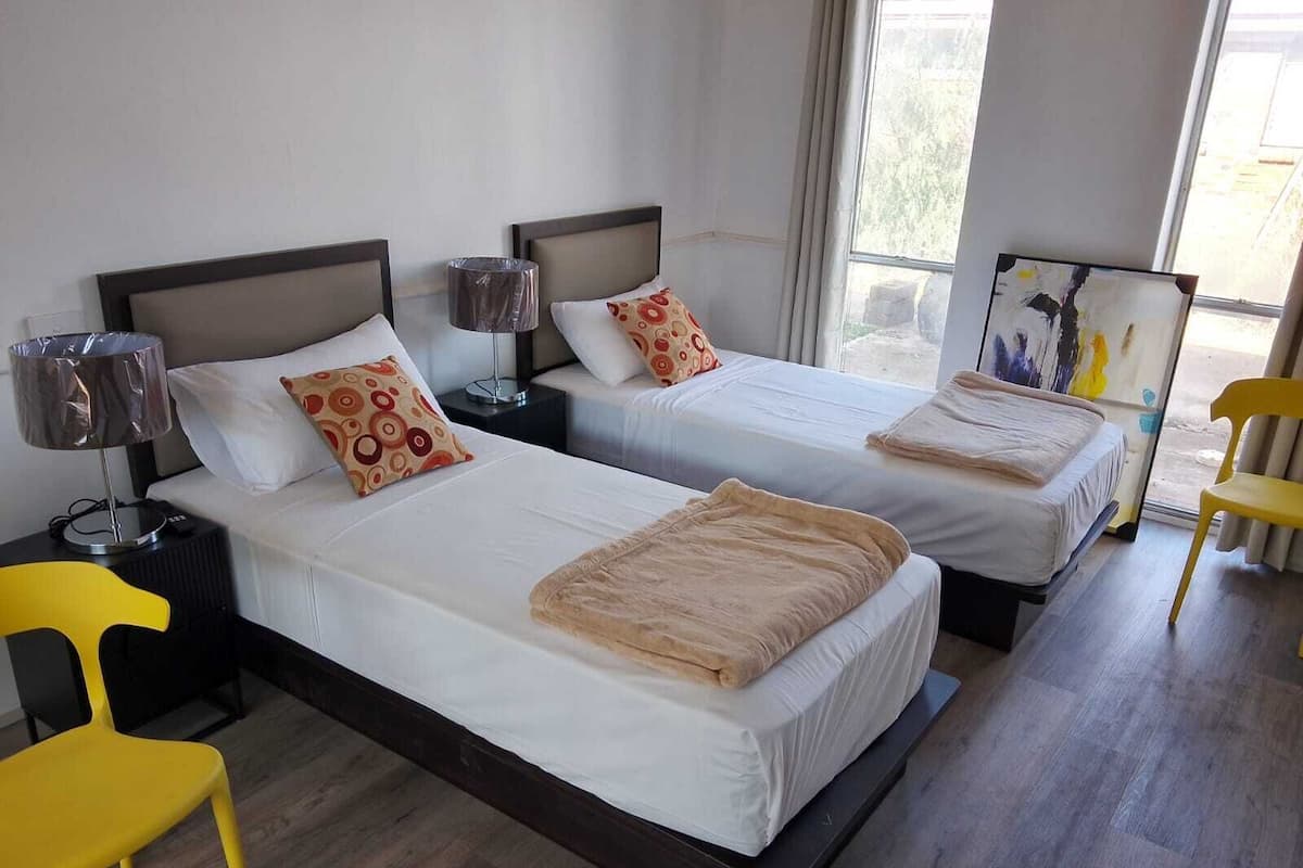 Une chambre d’hôtel avec deux lits, chacun étant équipé d’un oreiller et d’une lampe. Il y a une chaise et un tableau au mur.