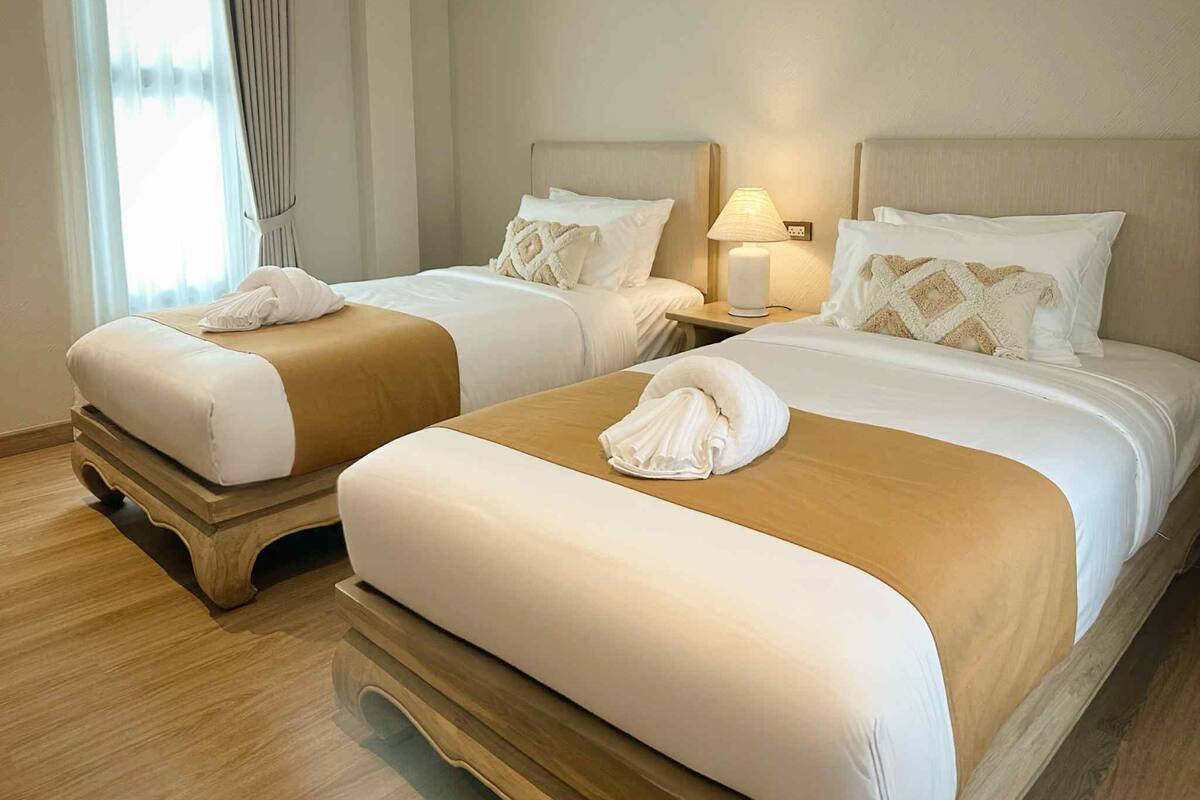 Dos camas con ropa de cama blanca y beige, una lámpara de noche y una ventana con cortinas.