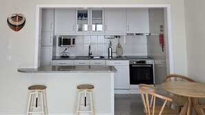 A modern kitchen with white cabinets, a microwave, a sink, and a stove. There are two stools and a wooden table with chairs. - Azulmaris - Directly on the beach | Terrace & sea view (Vila do Conde)
