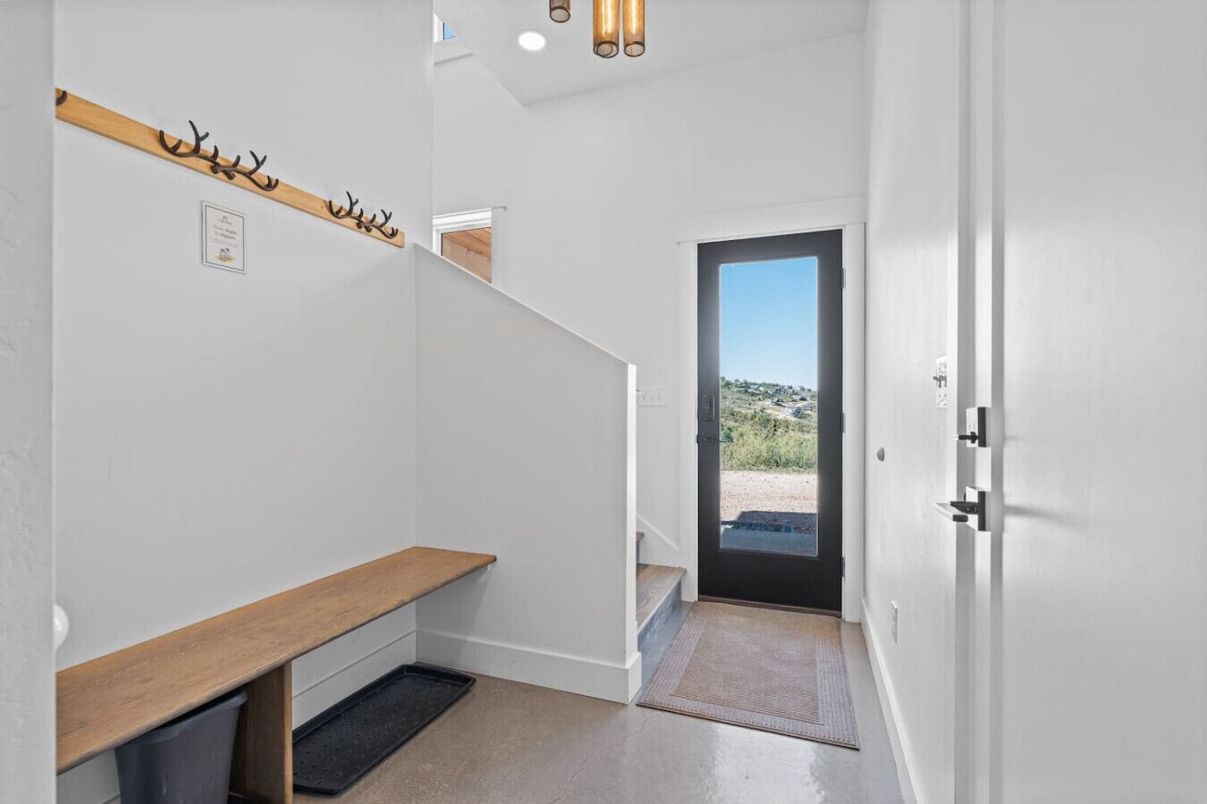 En moderne gang med en træbænk, knagerække og en dør, der fører til en balkon med udsigt over landskabet.