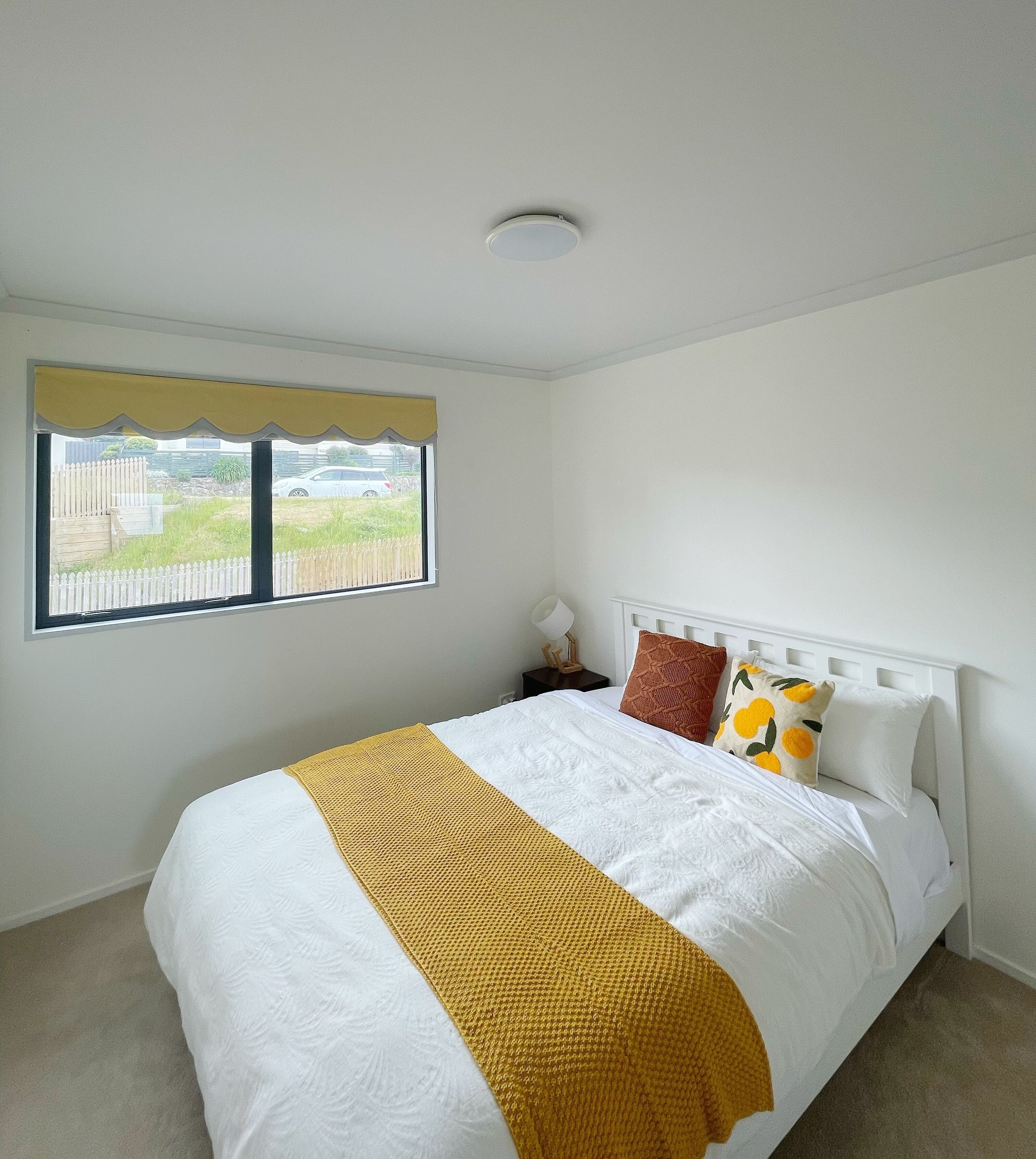 A bedroom with a bed, a window with a view of a fence and cars, and a ceiling light.