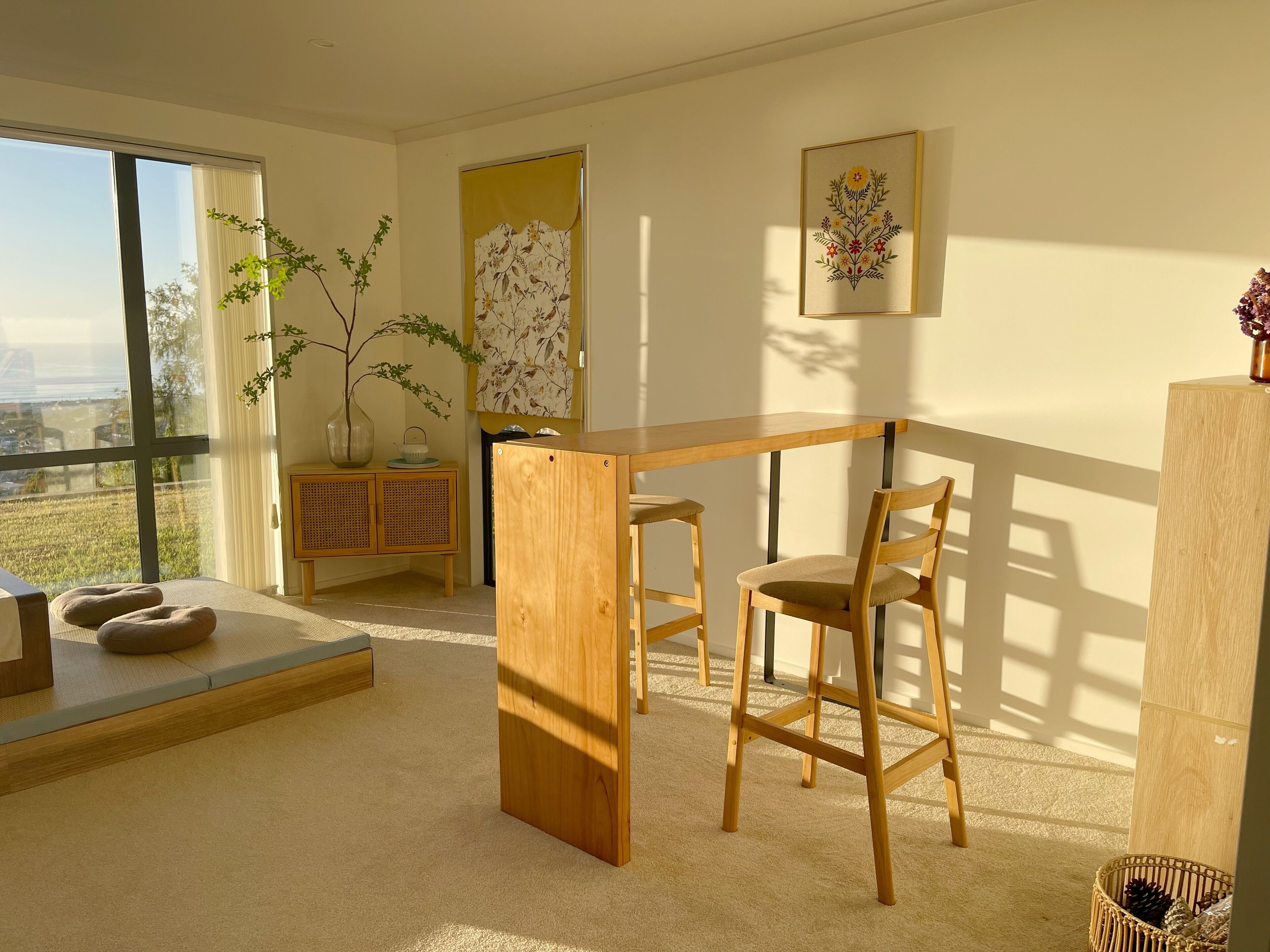 Een moderne kamer met een groot raam, een houten bureau, twee stoelen, een gestoffeerde bank en een rotan kastje, met een bloemig kunstwerk in een lijst aan de muur.