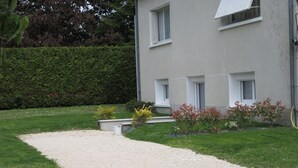 Ein Kiesweg führt zu einem Gebäude mit zahlreichen Fenstern, im Hintergrund eine Hecke.