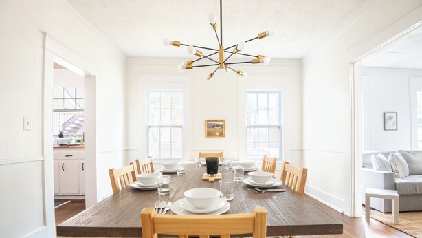 A dining room with a wooden table set for six, surrounded by wooden chairs. A modern chandelier hangs from the ceiling, and there are large windows on the walls. - Tri-City Traditions – Groups/WiFi/Downtown (Birmingham)