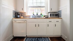 A kitchen with white cabinets, a dishwasher, a coffee maker, a microwave, and a window with plants. - Tri-City Traditions – Groups/WiFi/Downtown (Birmingham)