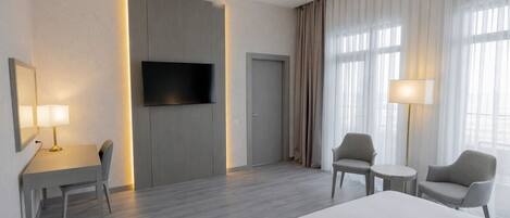 A hotel room with a bed, a desk with a chair, a TV mounted on the wall, and two chairs.