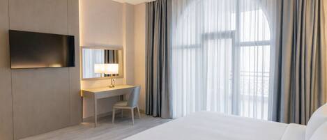 A hotel room with a bed, a desk with a chair, a TV mounted on the wall, and a large window with curtains.