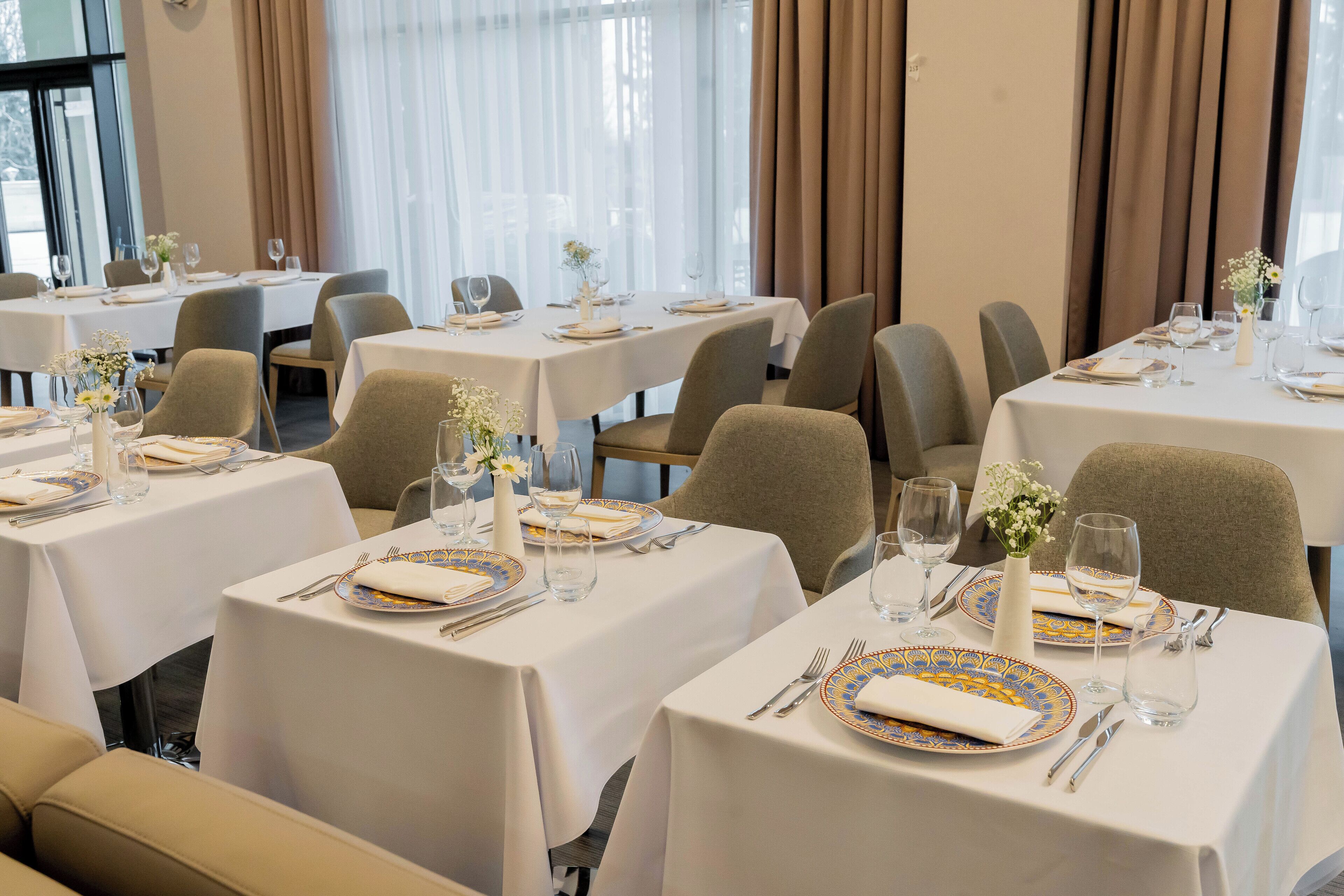 A dining area with multiple tables set for service, each with a white tablecloth, chairs, and table settings including plates, cutlery, and glassware.