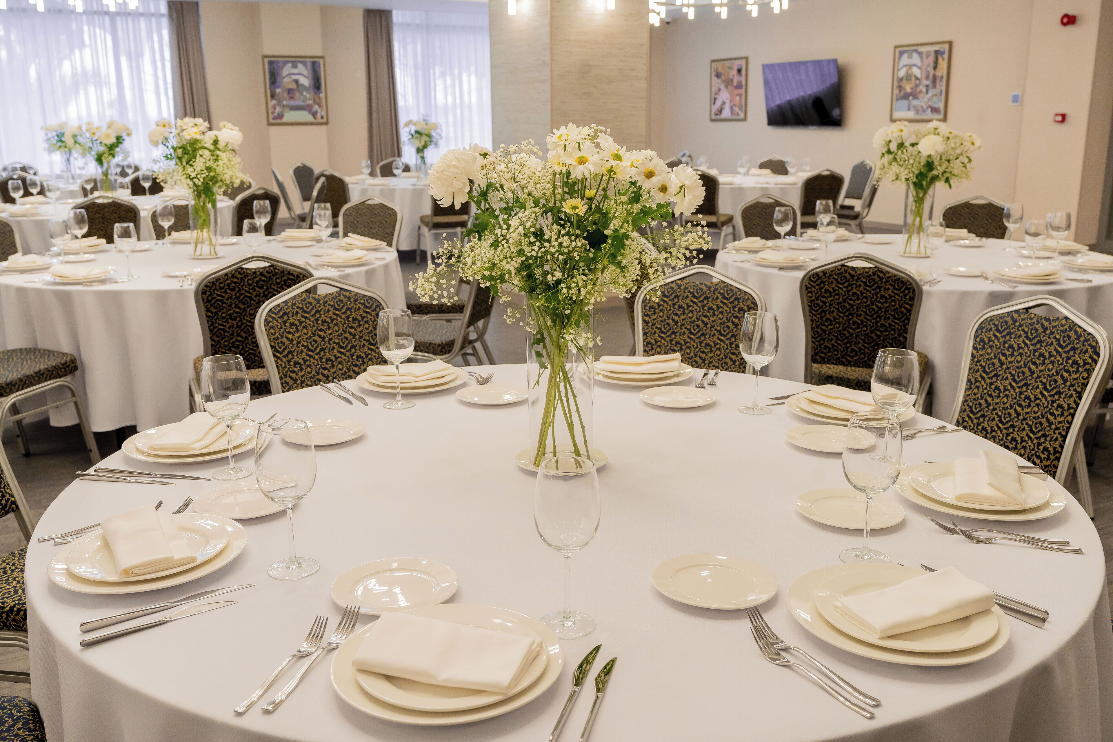 Un salón de banquetes con mesas redondas, cada una con mantel blanco, platos, cubiertos y centros de mesa florales.
