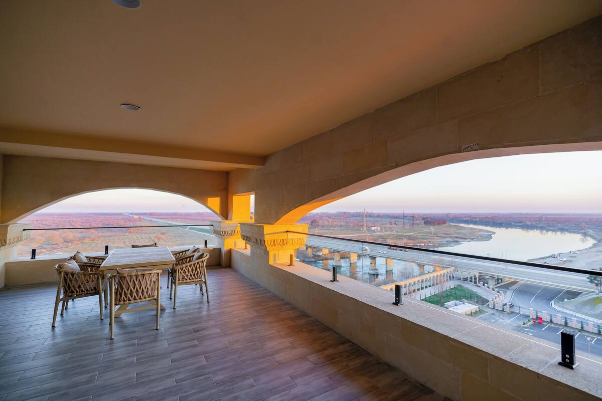 Een balkon met uitzicht op een rivier en een brug, met houten meubels en een stenen muur.
