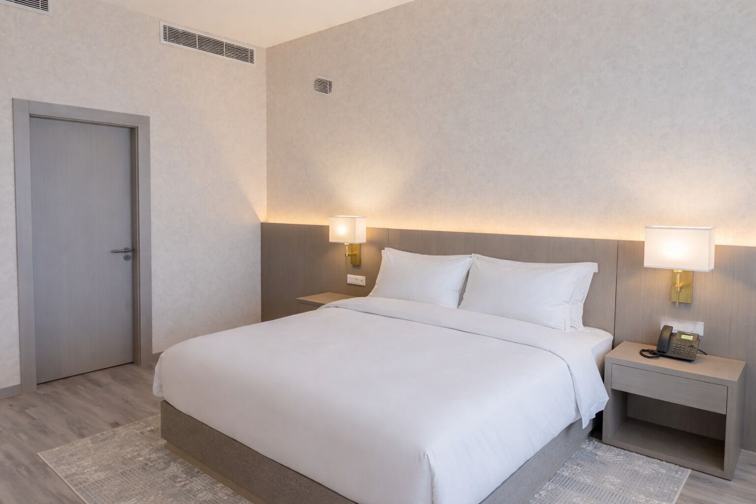 A hotel room with a bed, two bedside tables with lamps, a door, and a vent on the wall.