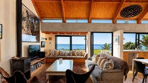 Un salon spacieux avec un grand canapé, une table basse et un téléviseur. De grandes fenêtres offrent une vue sur l’océan.