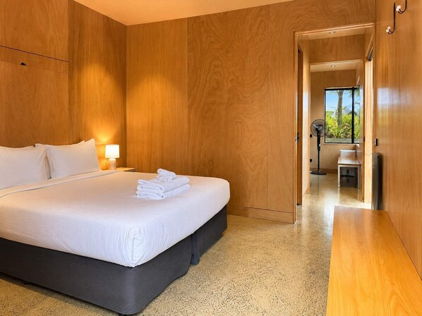 A modern hotel room with a large bed, wooden walls, a bedside table with a lamp, and a view through an open door.