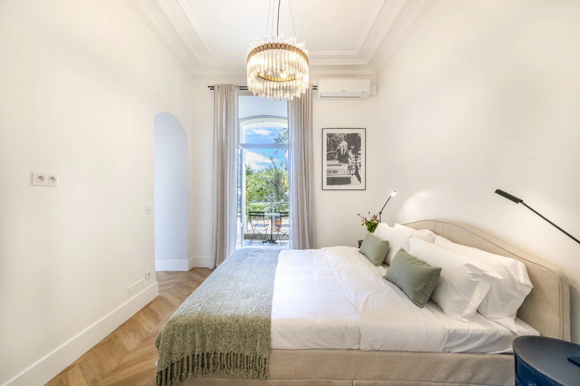Une chambre à coucher avec un lit, un lustre, un tableau au mur et une vue sur l’extérieur.