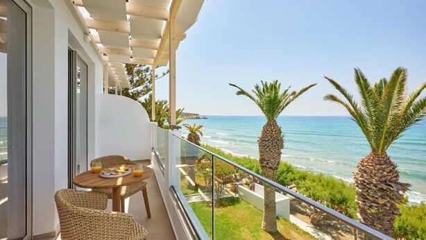 A balcony with a table and chairs, overlooking a beach with palm trees and the sea. - Artemis Hotel (Makry Gialos)