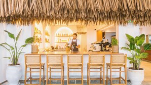 A bar area with a thatched roof, wooden bar stools, and potted plants. - Artemis Hotel (Makry Gialos)