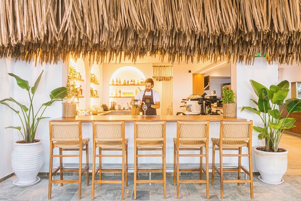 A bar area with a thatched roof, wooden bar stools, and potted plants. - Artemis Hotel (Makry Gialos)