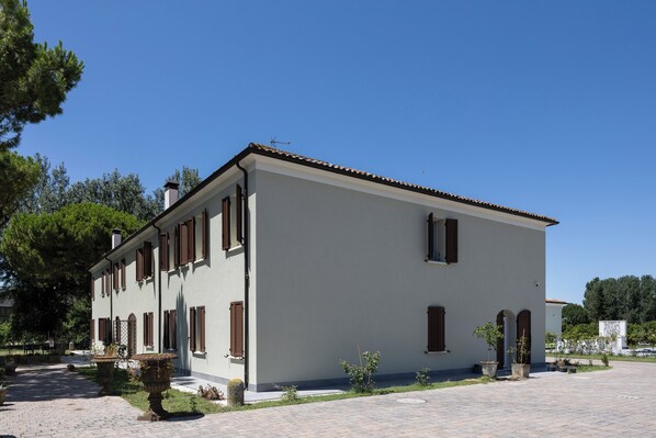 A two-story building with a white facade and brown shutters, surrounded by a paved courtyard with potted plants. - I 2 Pini Room&Breakfast (Portomaggiore)