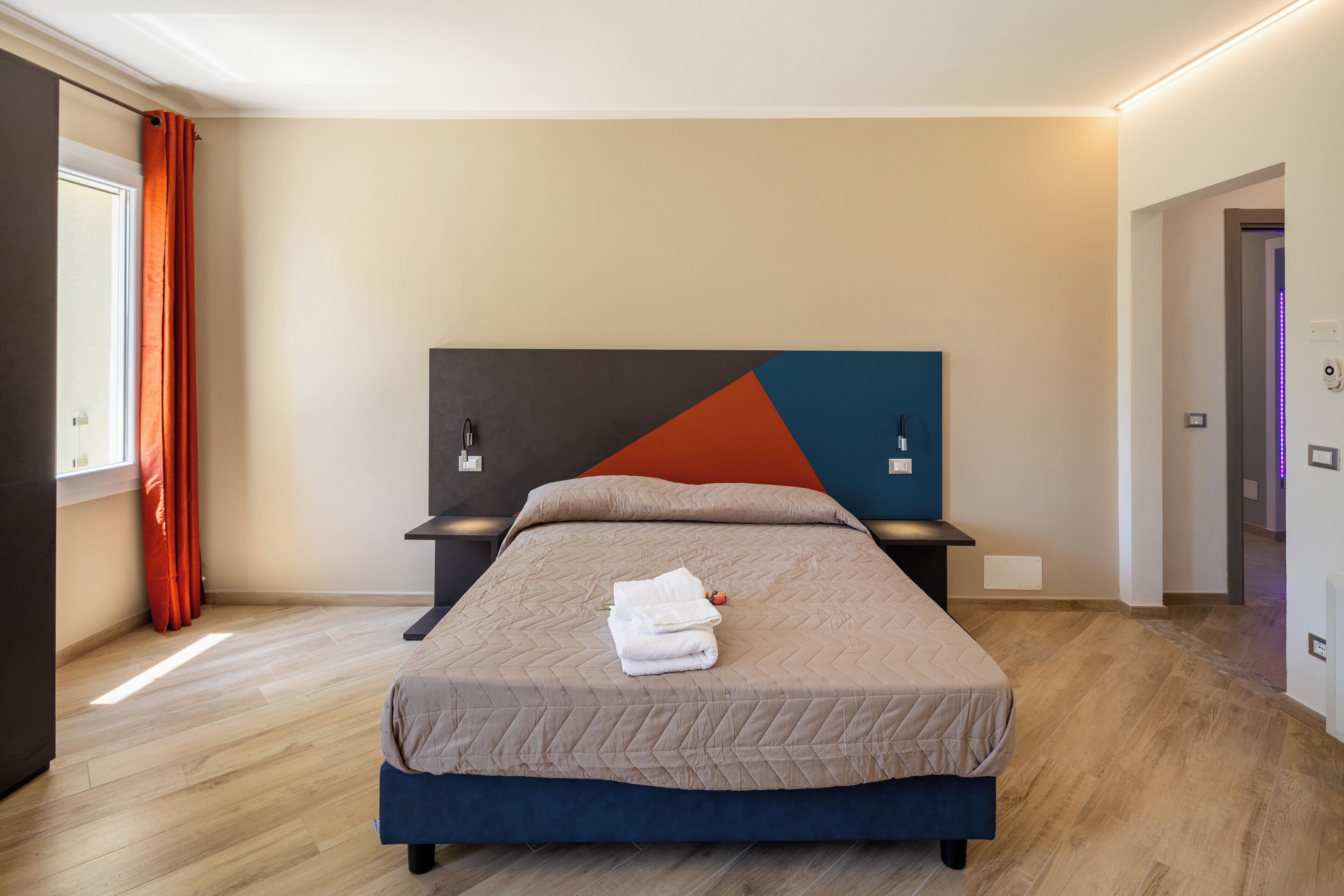 A modern hotel room with a large bed, a geometric wall art piece, and a view of the hallway.