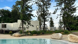 Zona de piscina con rocas grandes, árboles y un edificio blanco con un acceso.