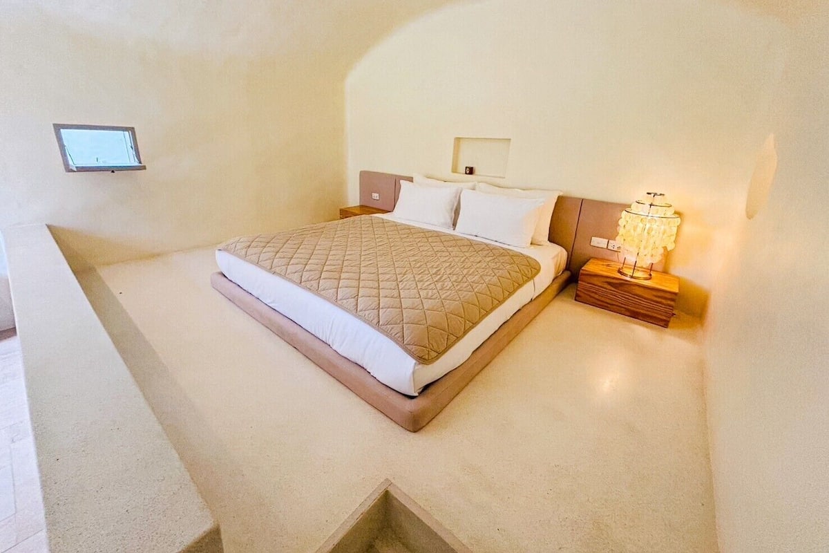 Un dormitorio con cama, mesitas de noche, una lámpara de pared y un ventanal pequeño.