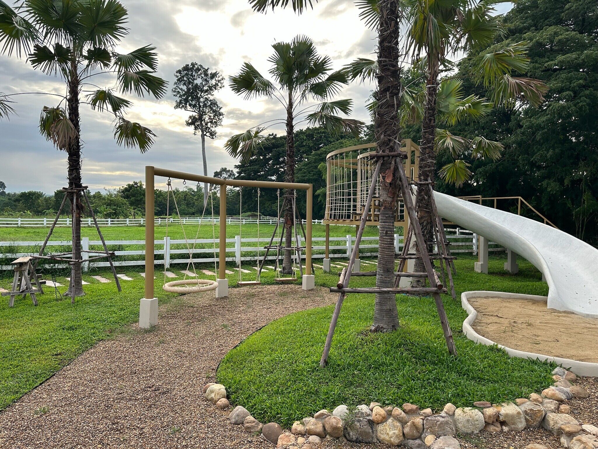 Un parque infantil con columpios, tobogán y estructura de escalada, rodeado de palmeras y una cerca blanca.