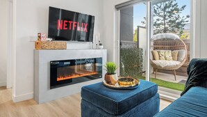 A modern living room with a fireplace, a flat-screen TV displaying the Netflix logo, a wicker basket, a potted plant, and a wicker chair with cushions. - ✨ The Era City Haven – Modern 2BR/2BA w/ Breathtaking Calgary Views (Calgary)