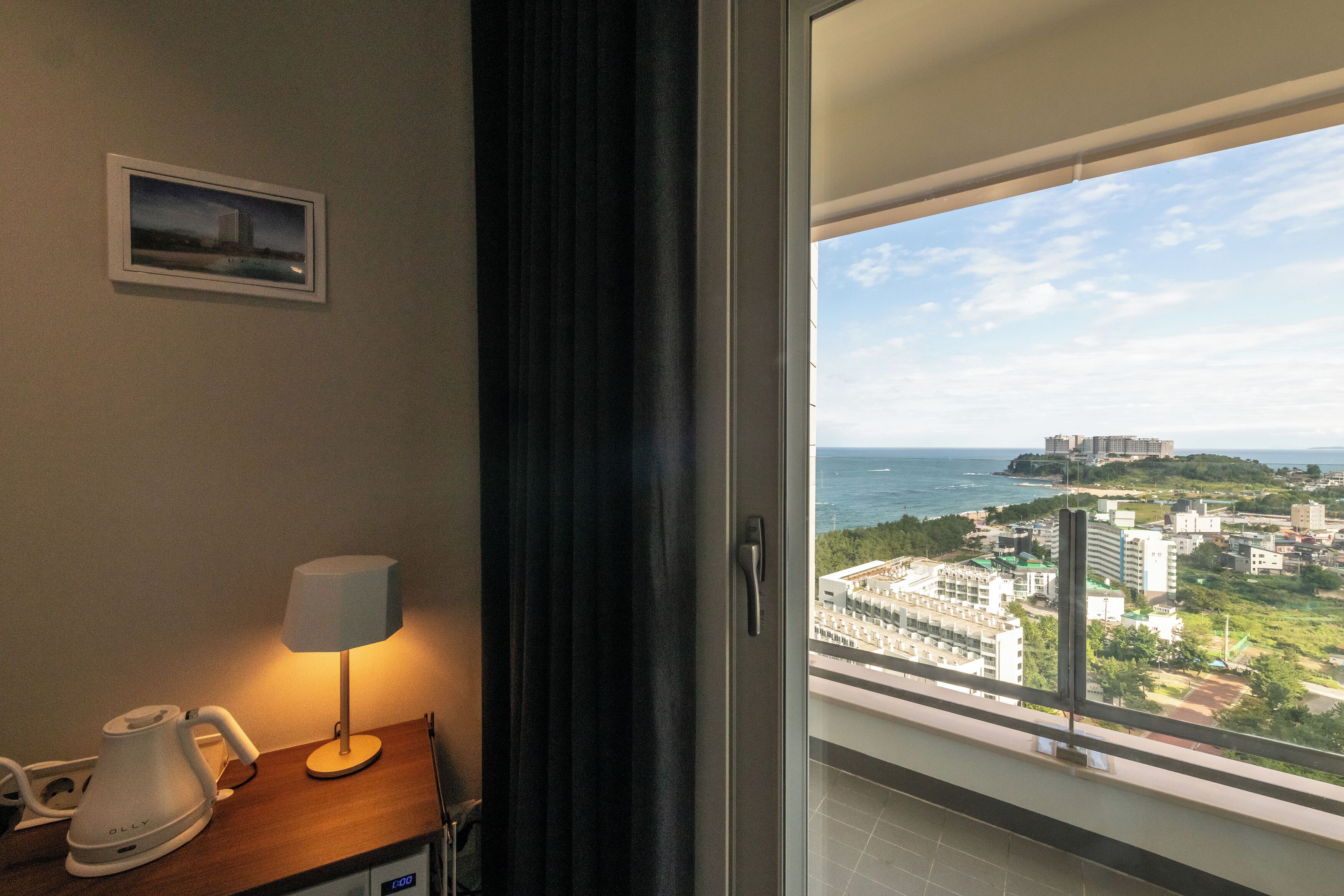 A hotel room with a view of a cityscape, a lamp, and a kettle on the table.