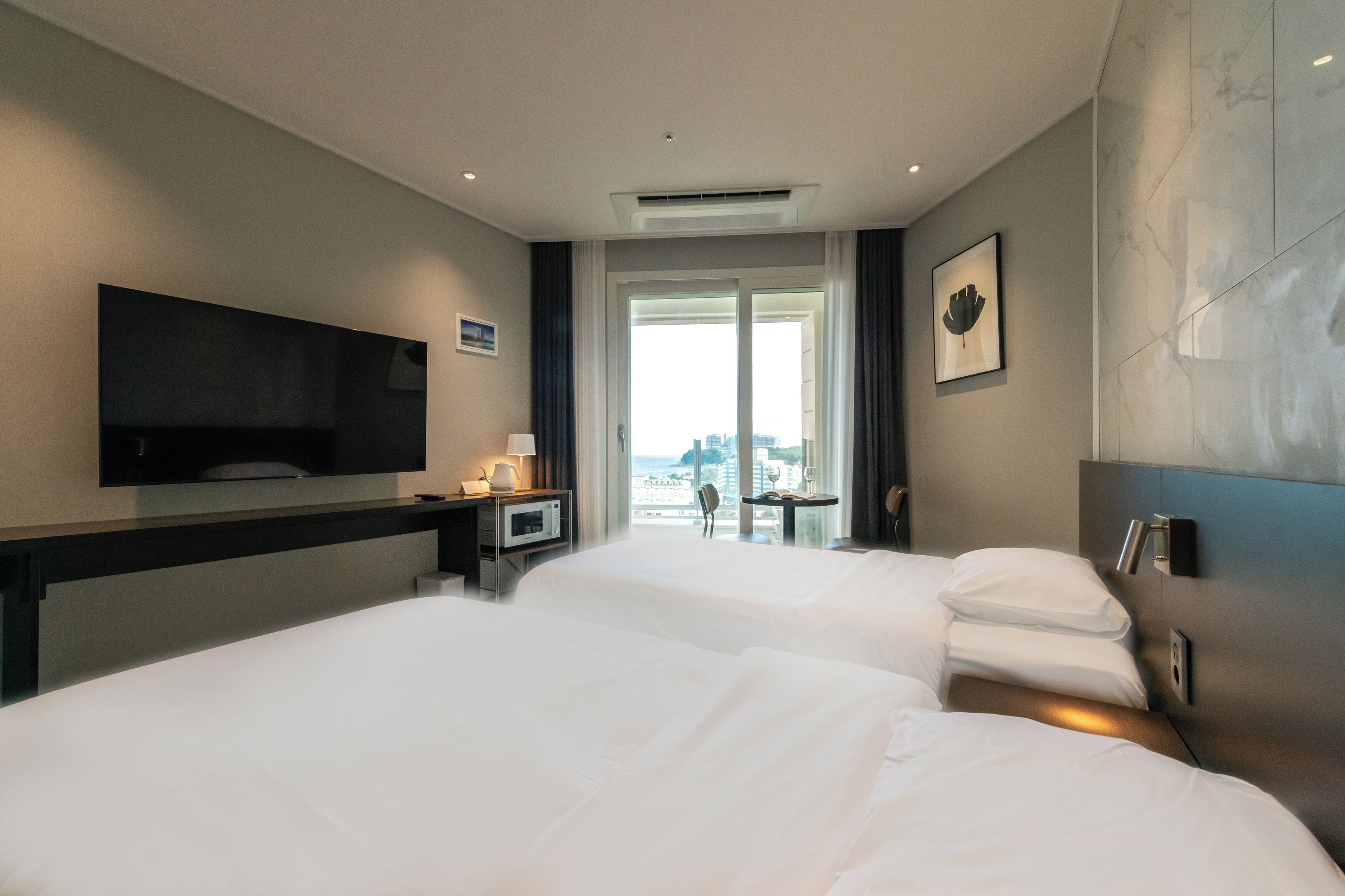 A hotel room with two beds, a flat-screen TV, a desk with a lamp, and a view of the city.