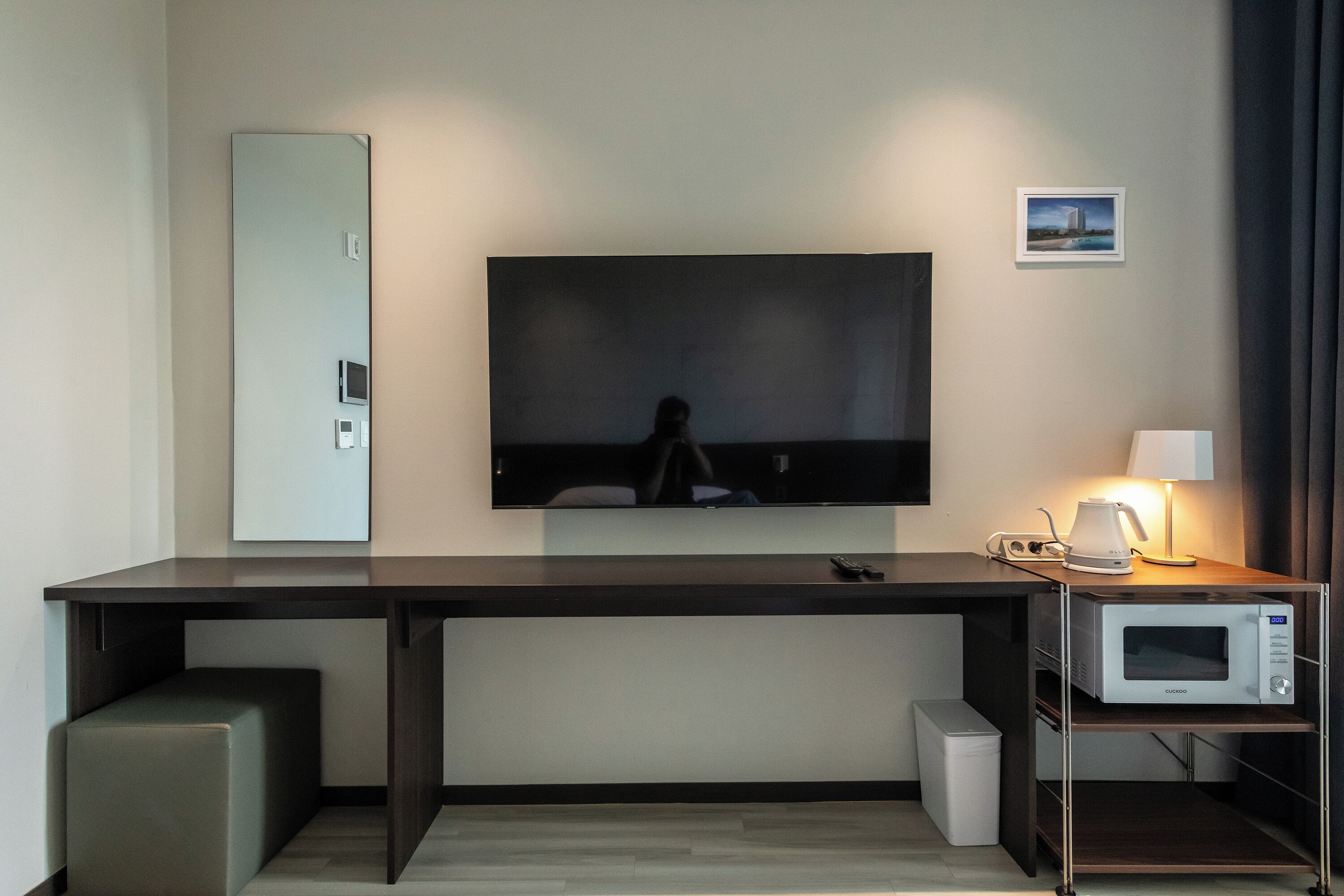 Ein modernes Hotelzimmer mit großem Flachbildfernseher an der Wand, einem Schreibtisch mit Mikrowelle und einem kleinen Nachttisch mit Lampe.