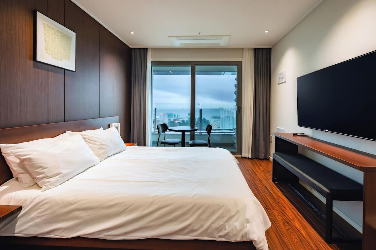 A hotel room with a large bed, a TV, a balcony with a table and chairs, and a city view.