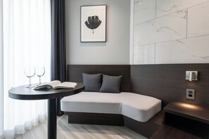 Ein modernes Hotelzimmer mit einem bogenförmigen Bett, einem runden Tisch mit Buch und Gläsern, einem gerahmten Kunstwerk an der Wand und einer Wand mit Marmoroptik.