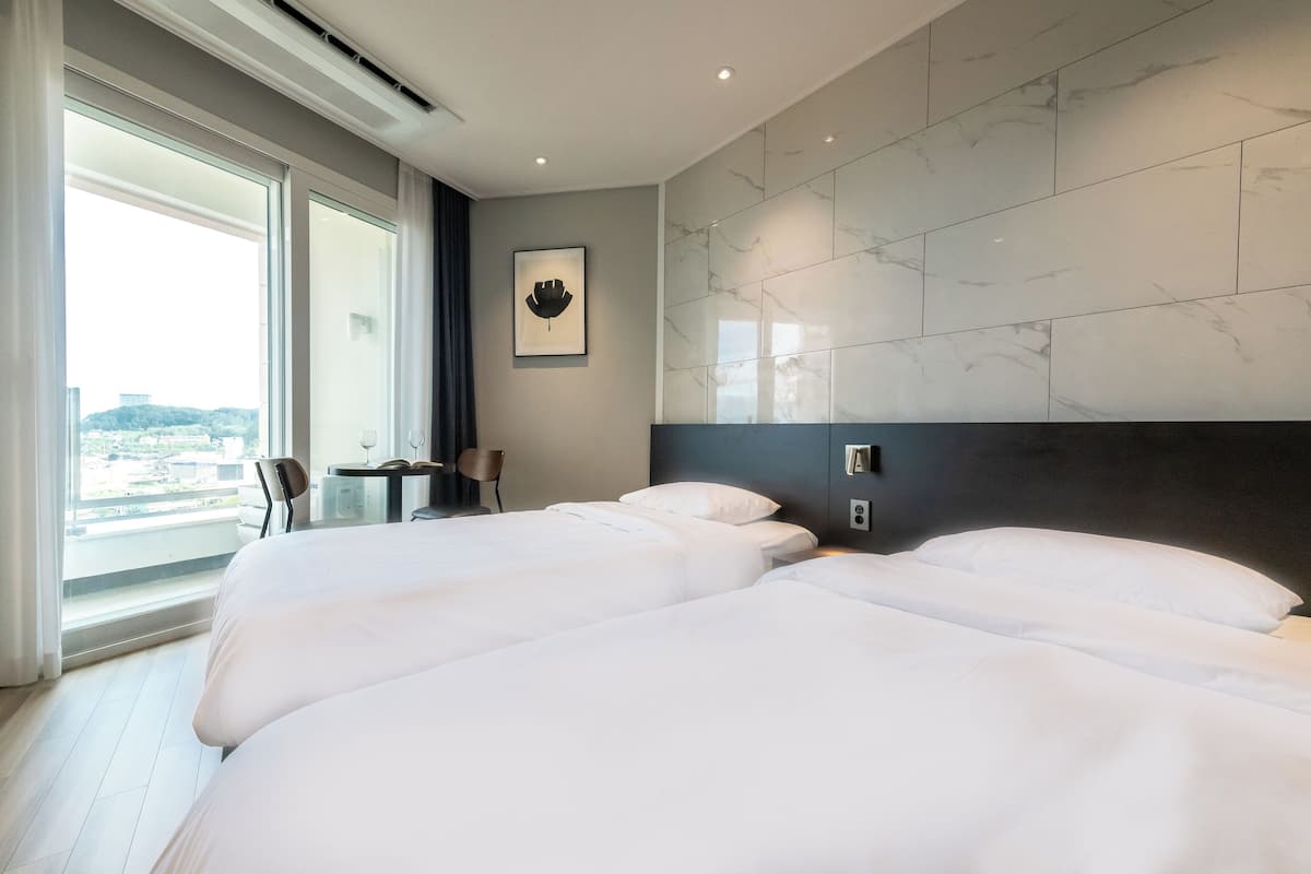 A hotel room with two beds, a large window with a city view, a small table, and a chair.