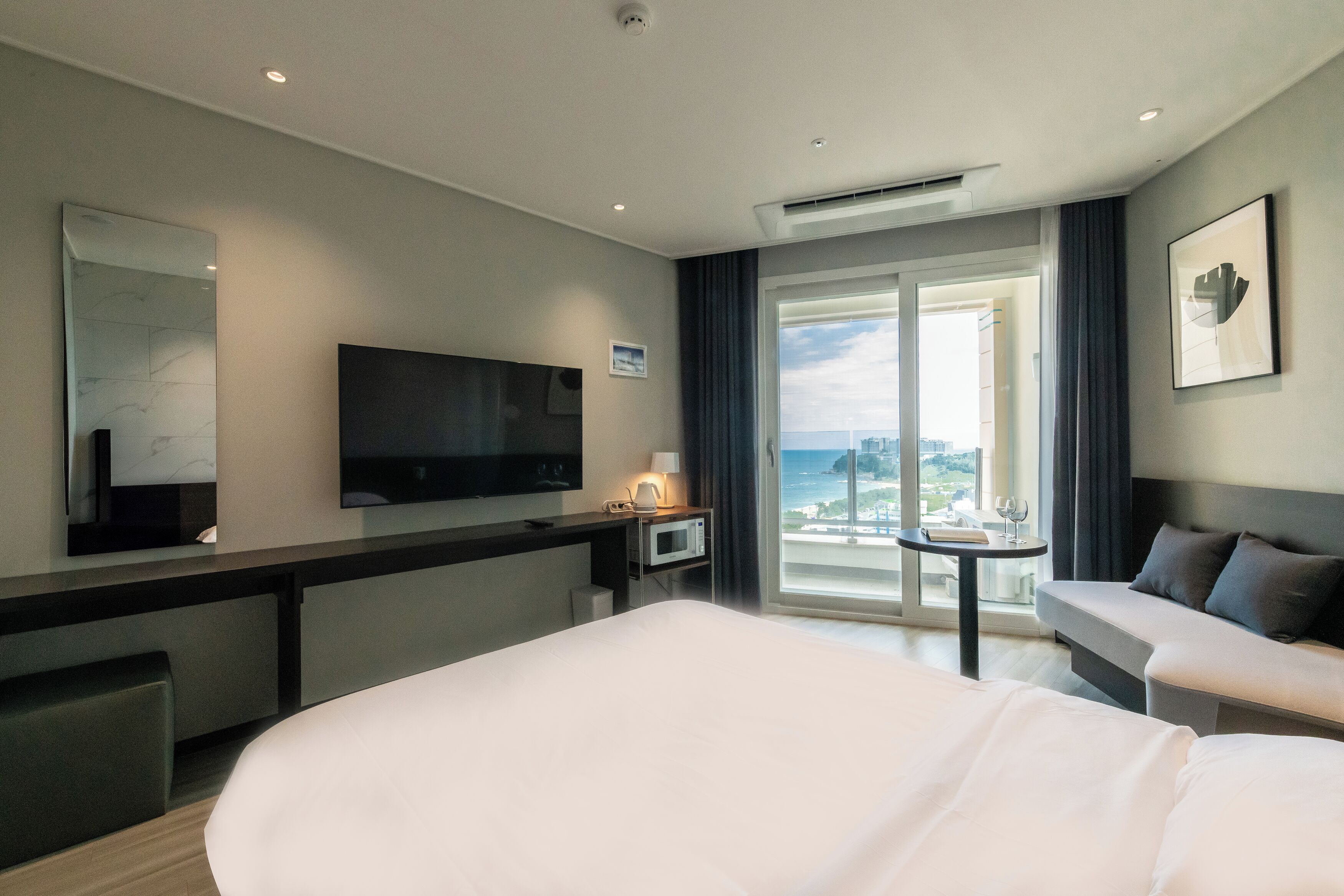 Habitación de hotel con una cama grande, un televisor de pantalla plana, un escritorio con lámpara, una mesita con una copa de vino, un sofá y un ventanal con vistas.