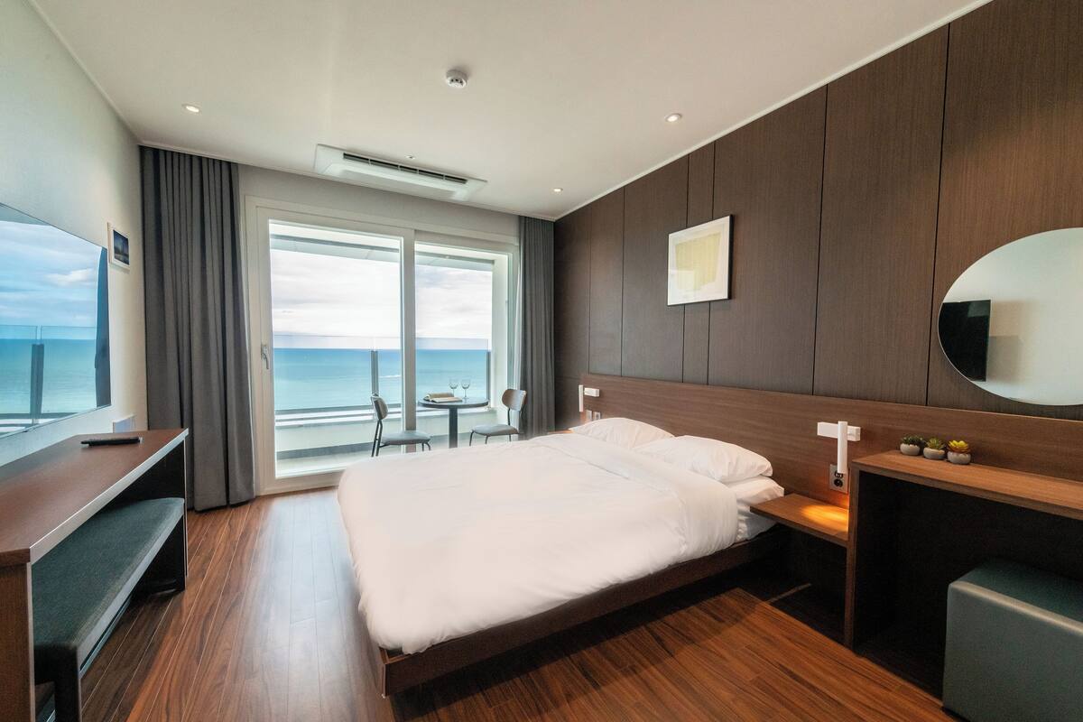 A modern hotel room with a large bed, a desk, a chair, and a small table. There is a large window with a view of the ocean.