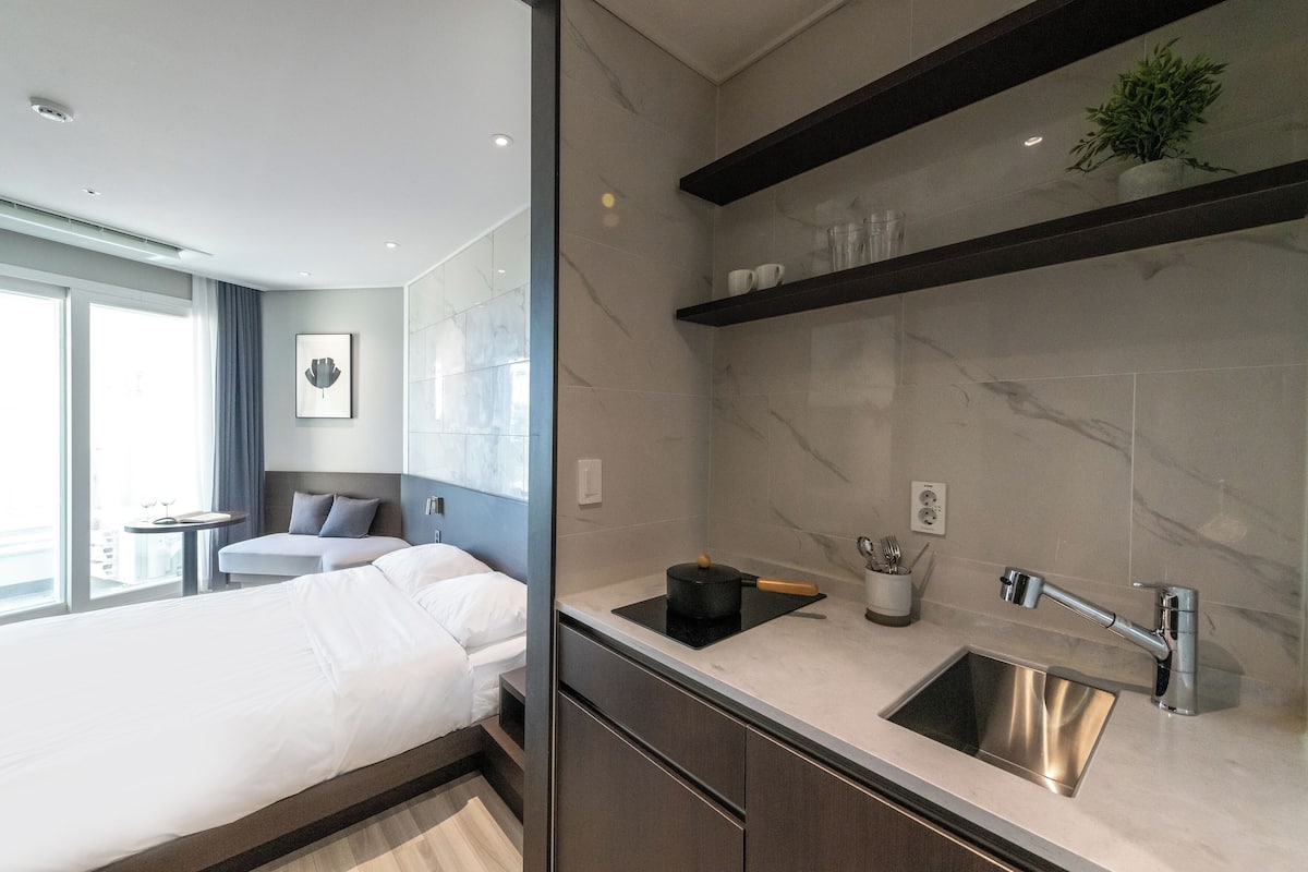 A modern hotel room with a bed, bedside table, and a kitchenette with a sink and countertop.