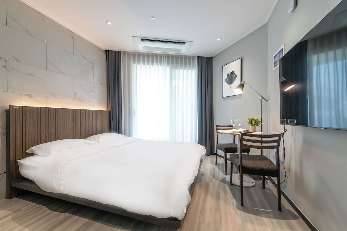 A modern hotel room with a large bed, a desk with a chair, a small table with a plant, and a large window with curtains.